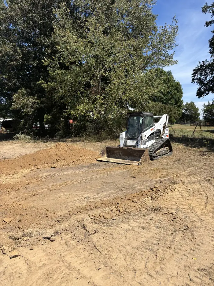 A skid steer with a loader bucket on a freshly cut dirt pad beside a spoil pile